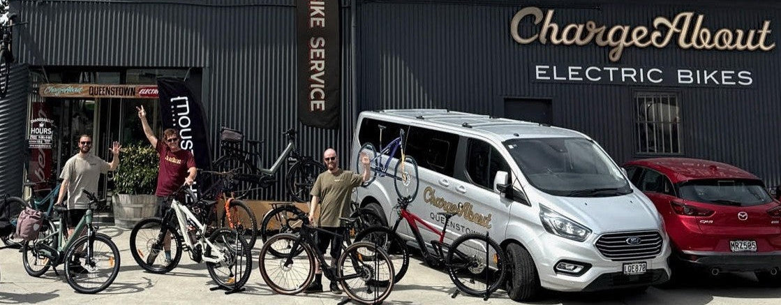 A group of people standing with electric bicycles in front of a dark gray building labeled 'ChargeAbout Electric Bikes', with a white branded van parked beside them and a red car on the right.