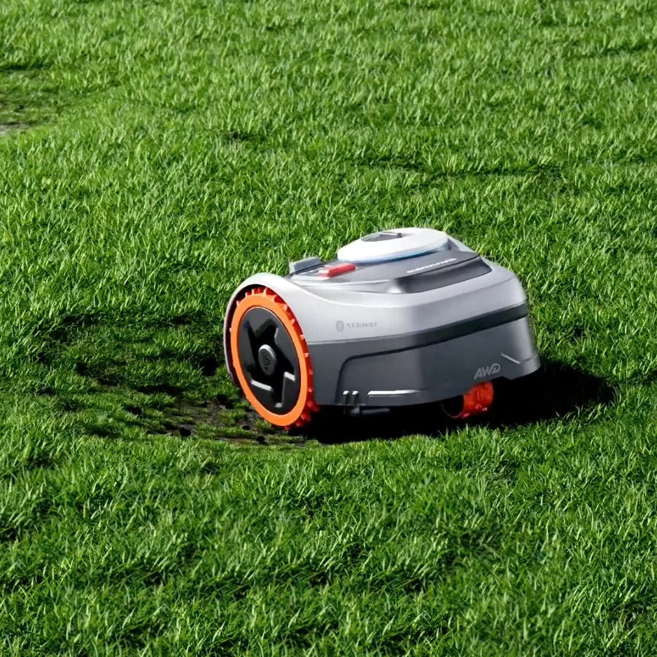 A robotic lawnmower with a gray body and bright orange wheel accents, shown operating on a green grass lawn.