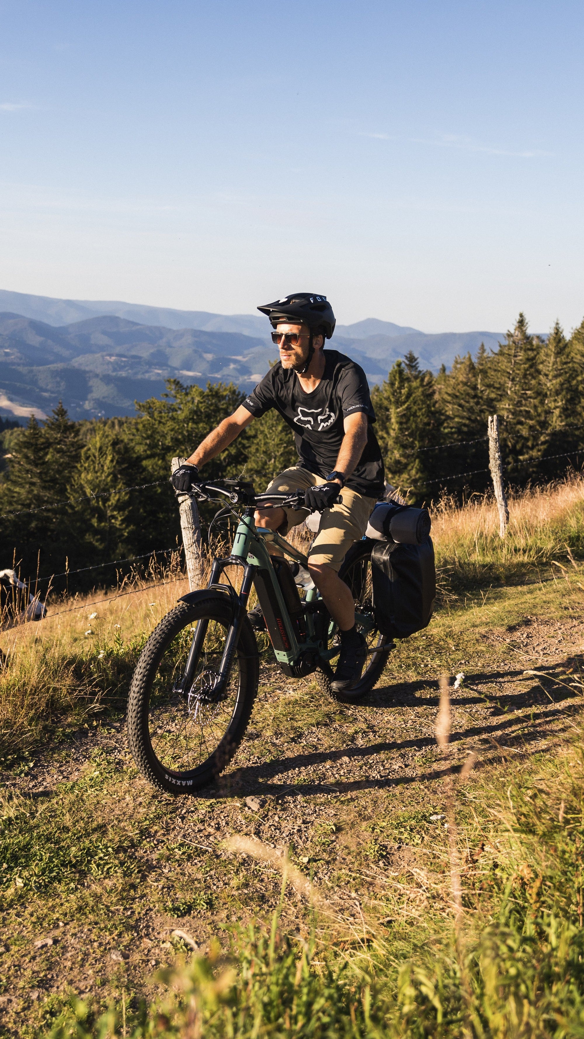 A man wearing a black helmet and sunglasses rides an electric mountain bike on a grassy dirt path with a scenic mountain and forest background.
